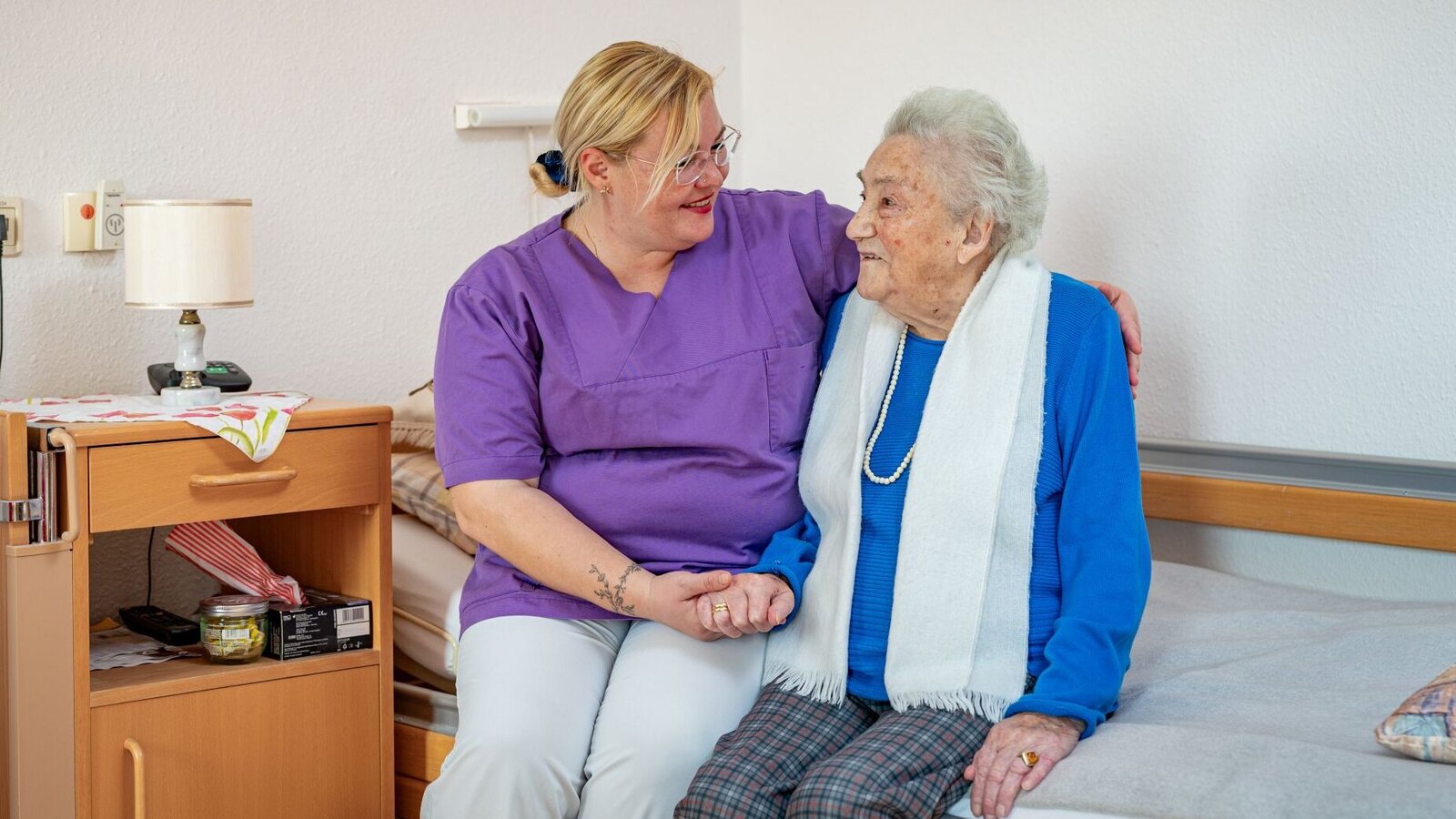 Pflegerin sitzt zusammen mit Bewohnerin auf dem Bett